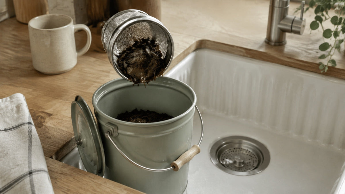 Cooled loose tea leaves emptied from a basket infuser into a food-waste caddy beside a sink strainer