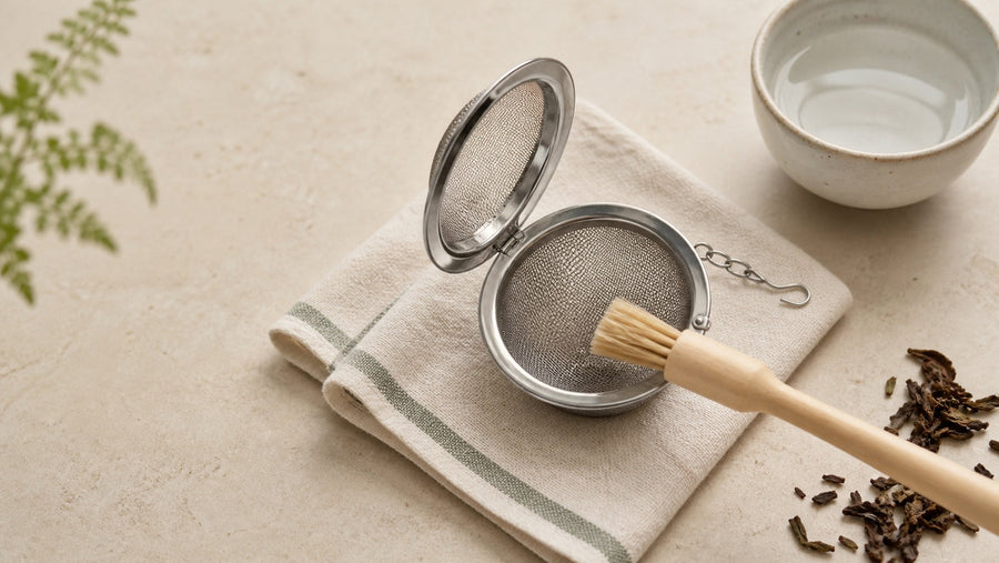 Stainless-steel tea infuser being cleaned with a soft brush beside a bowl of water